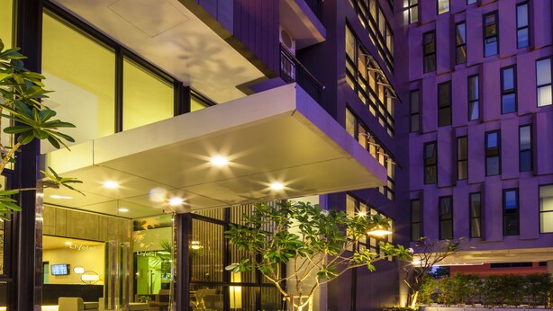 A modern hotel entrance with a glass facade, outdoor seating, and a parking area.
