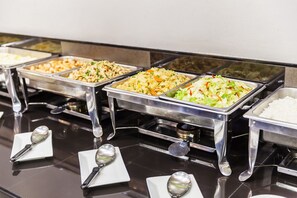 Un buffet proposant une variété de plats, notamment des salades, du riz et d’autres mets, servis dans des plateaux en métal avec des cuillères sur des assiettes blanches.