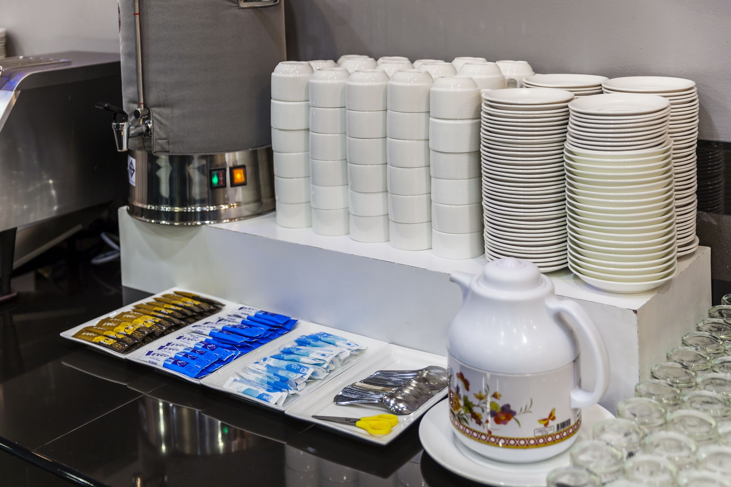 Een buffetopstelling met stapels witte borden, een koffieapparaat en een schaal met maaltijdverpakkingen.