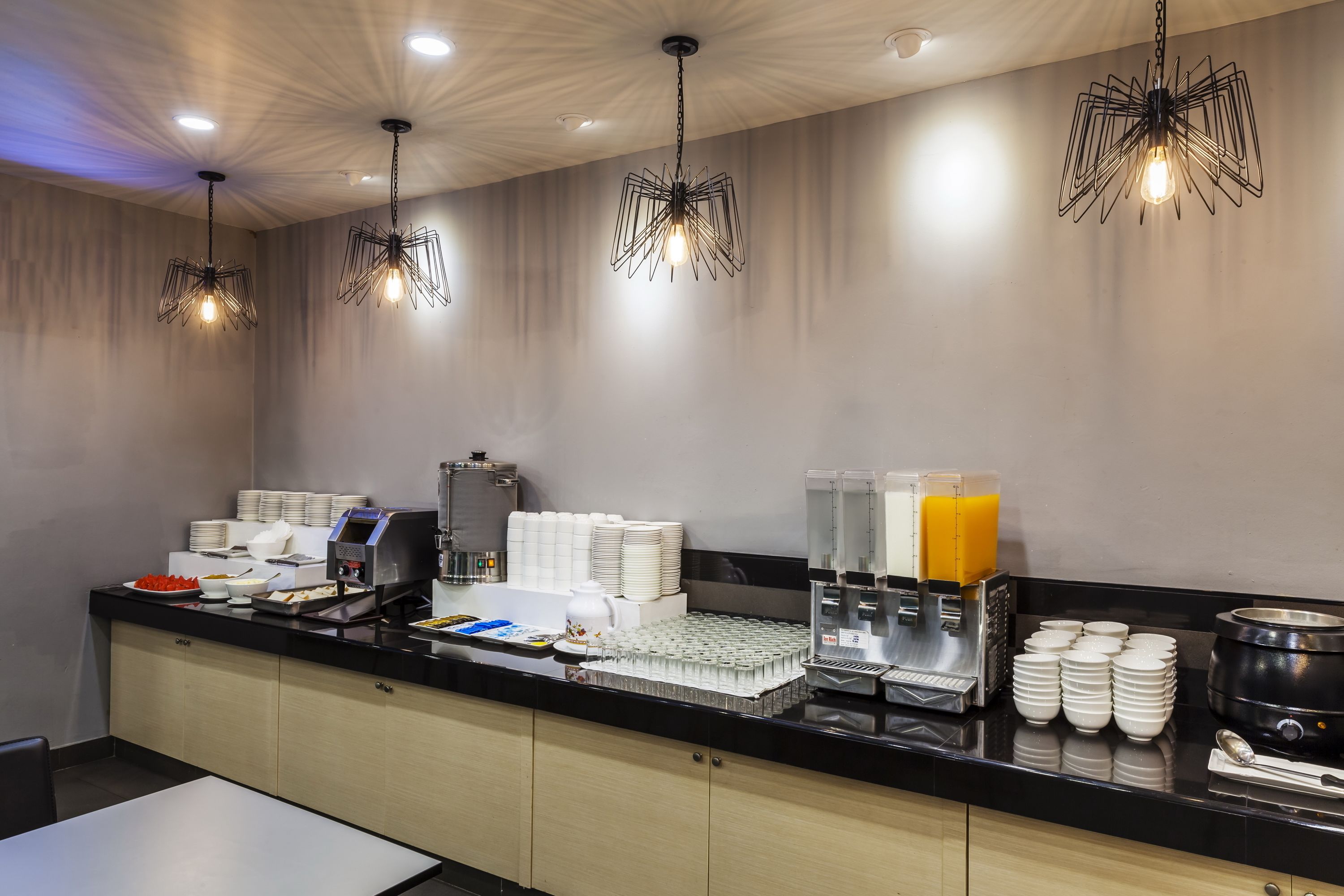 Zona de desayuno de hotel con barra de buffet, que incluye platos, tazones y una máquina de café.