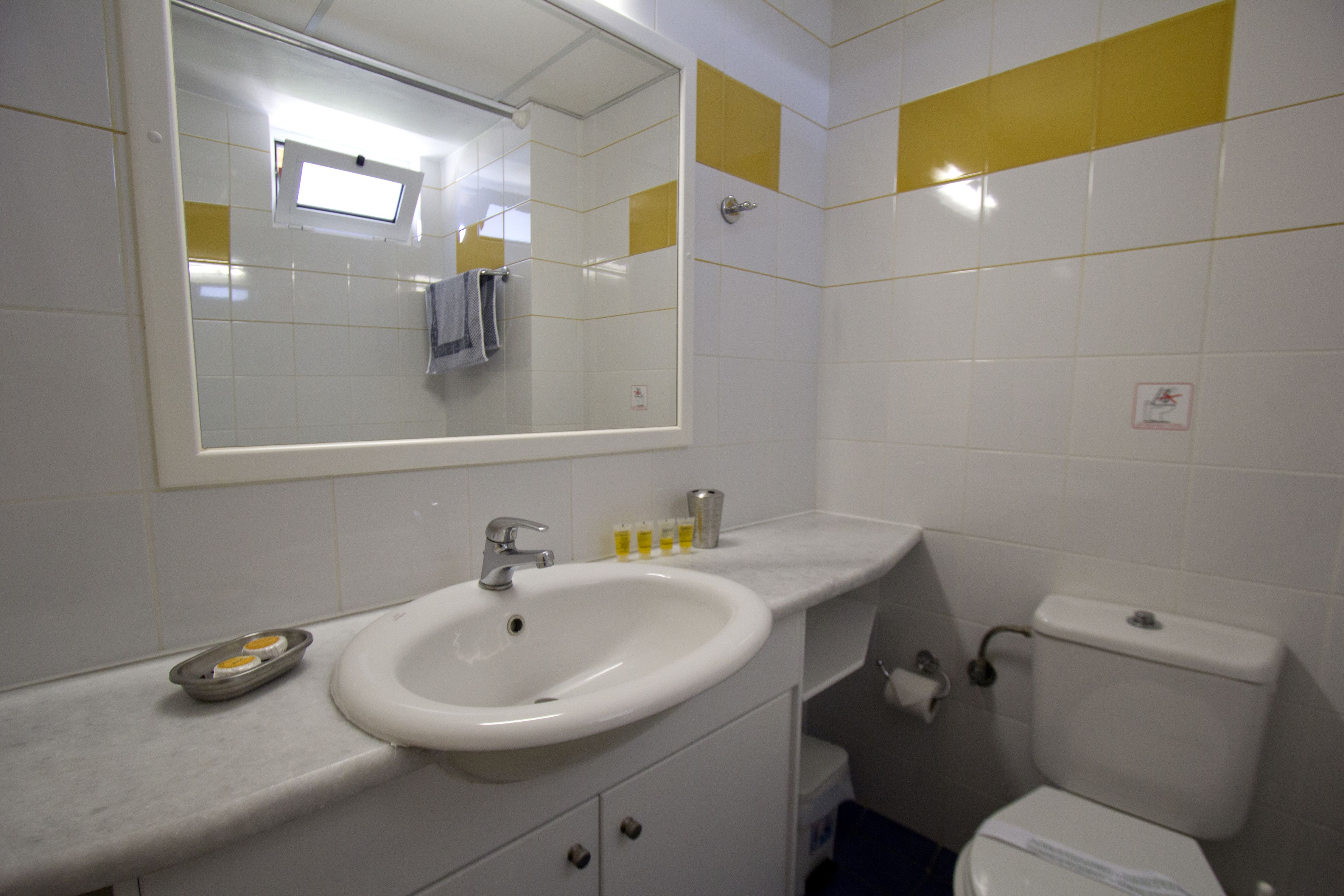 Un baño con lavamanos blanco, espejo e inodoro. Las paredes tienen azulejos amarillos y hay una ventana sobre el lavamanos.