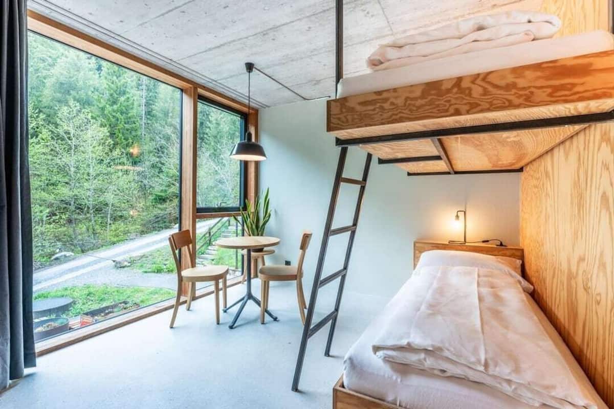 A modern room with a bunk bed, a small table with chairs, a potted plant, and a view of a forest through large windows.