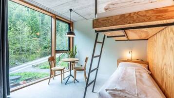 A modern room with a bunk bed, a small table with chairs, a potted plant, and a view of a forest through large windows.
