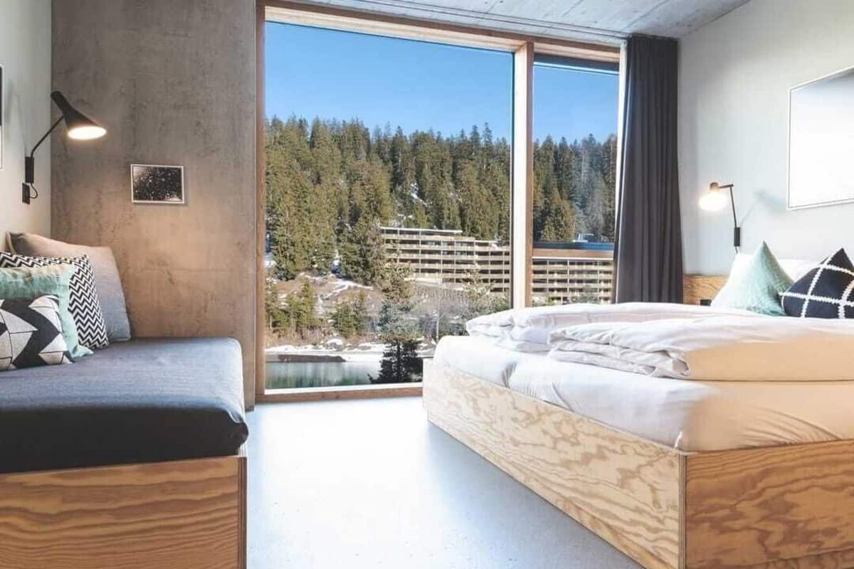 A modern bedroom with a large window, a bed, a sofa, and a view of a forest and buildings.