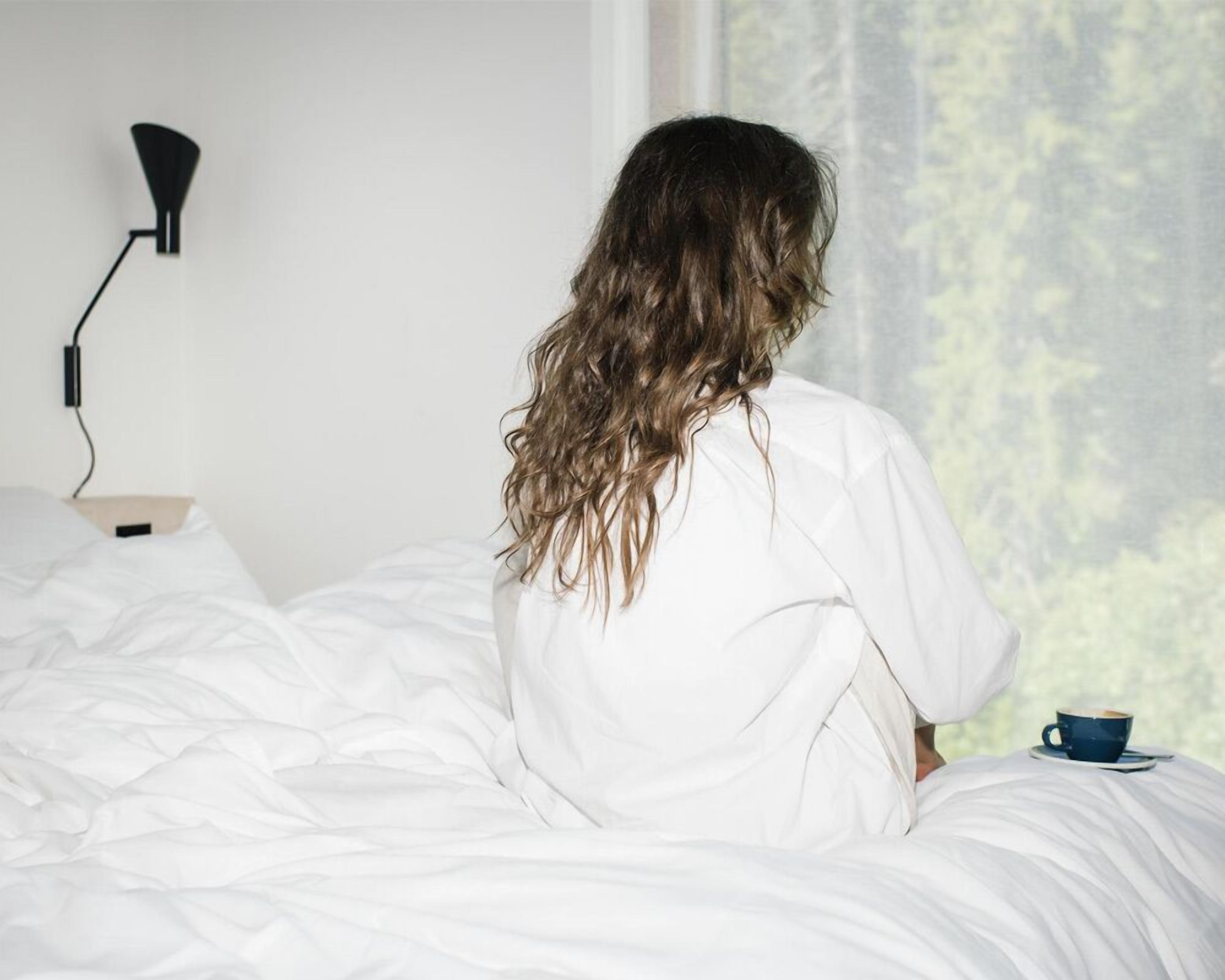 A person sitting on a bed in a room with a window, a lamp, and a cup on the bedside table.