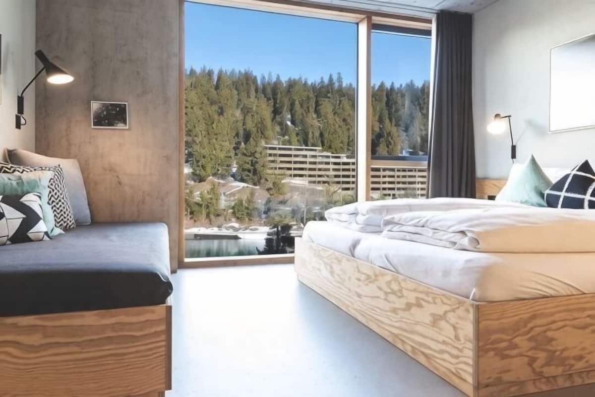 A modern hotel room with a large bed, a sofa, and a view of a forest and buildings.