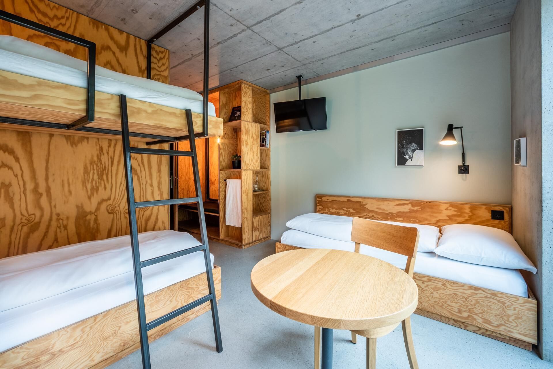 A compact room with bunk beds, a round wooden table, a TV mounted on the wall, and a small shelf with books and a plant.