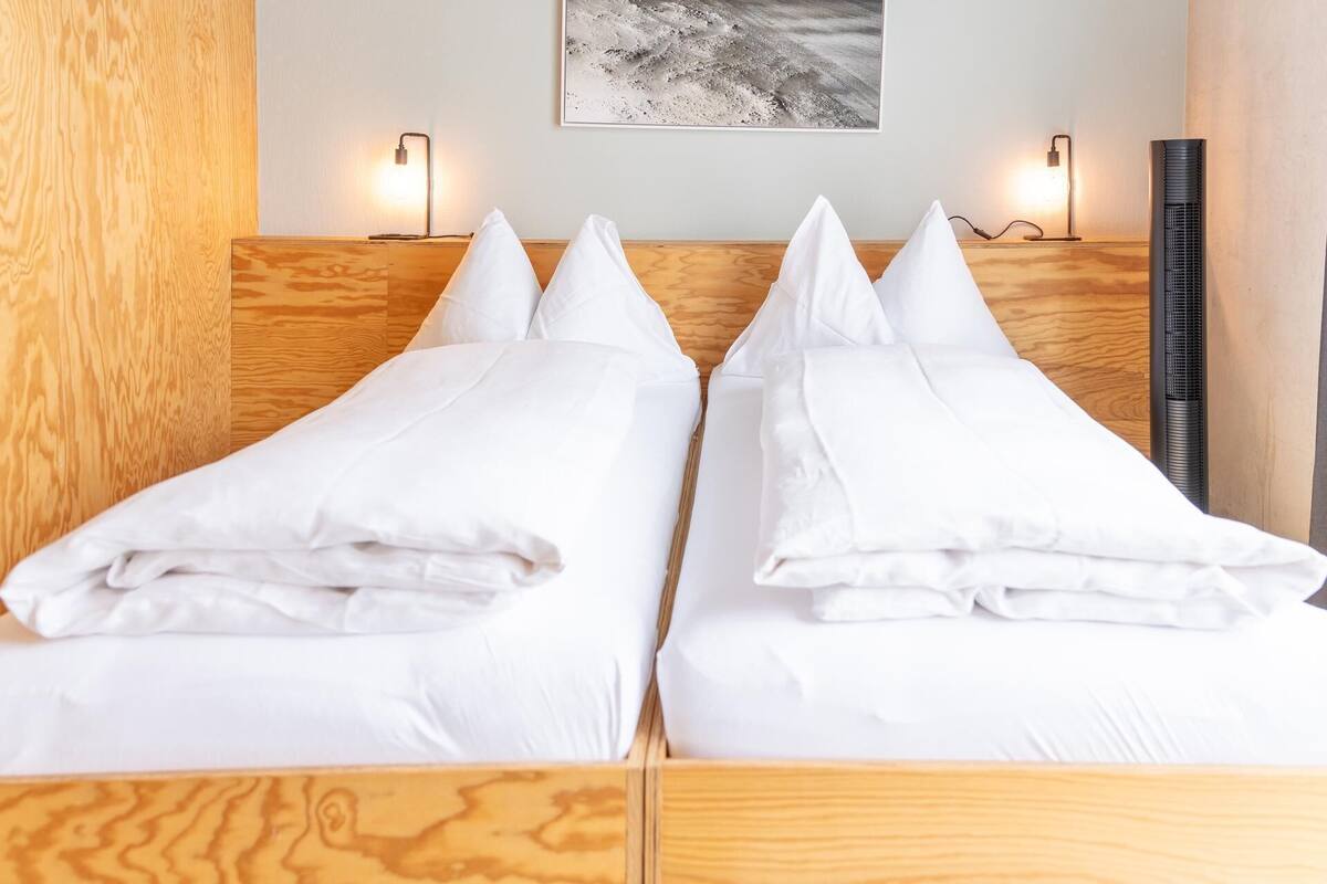 A double bed with white linens and pillows, a wooden headboard, two wall-mounted lamps, and a framed picture above the bed.