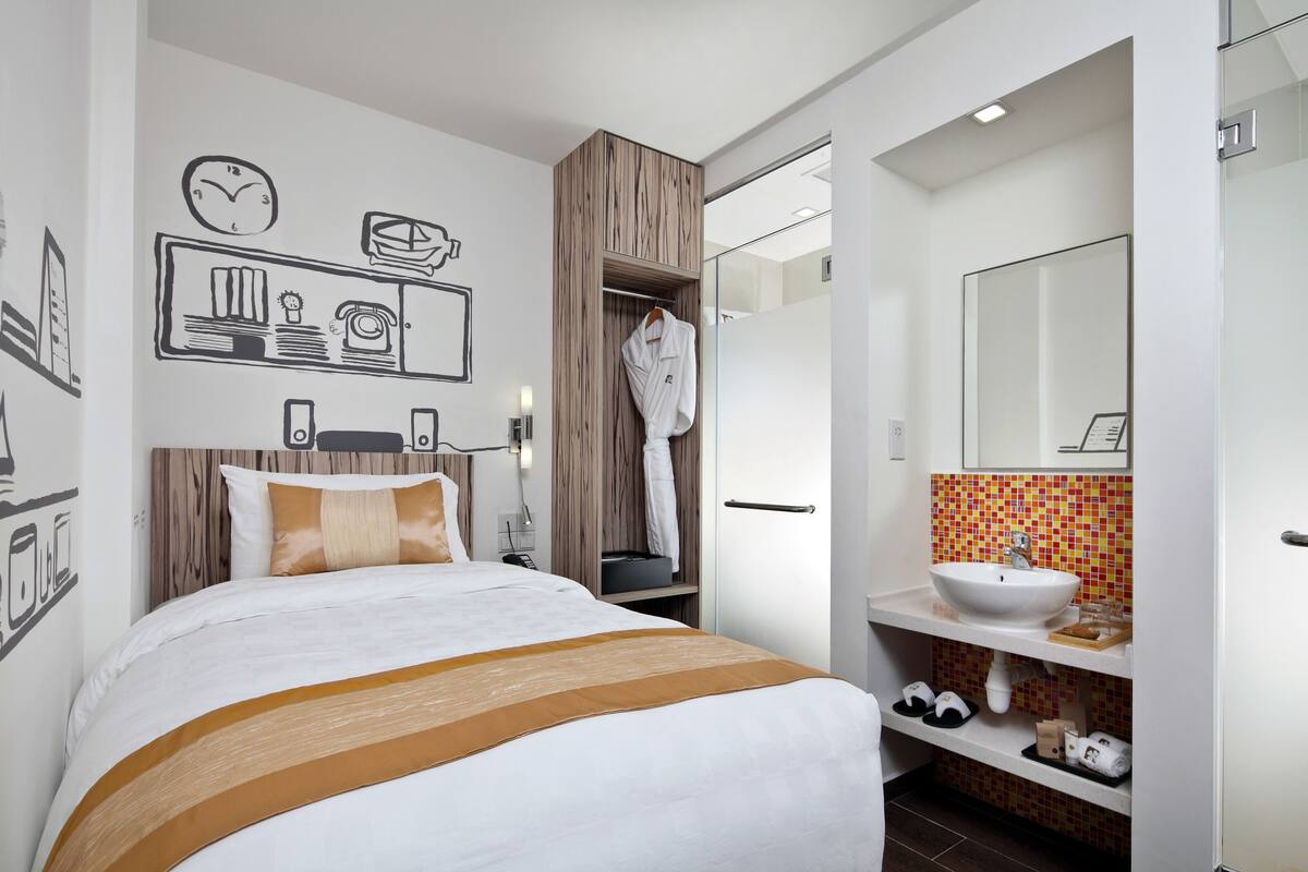 A modern hotel room with a bed, a wardrobe, a bathroom with a sink and mirror, and wall art.