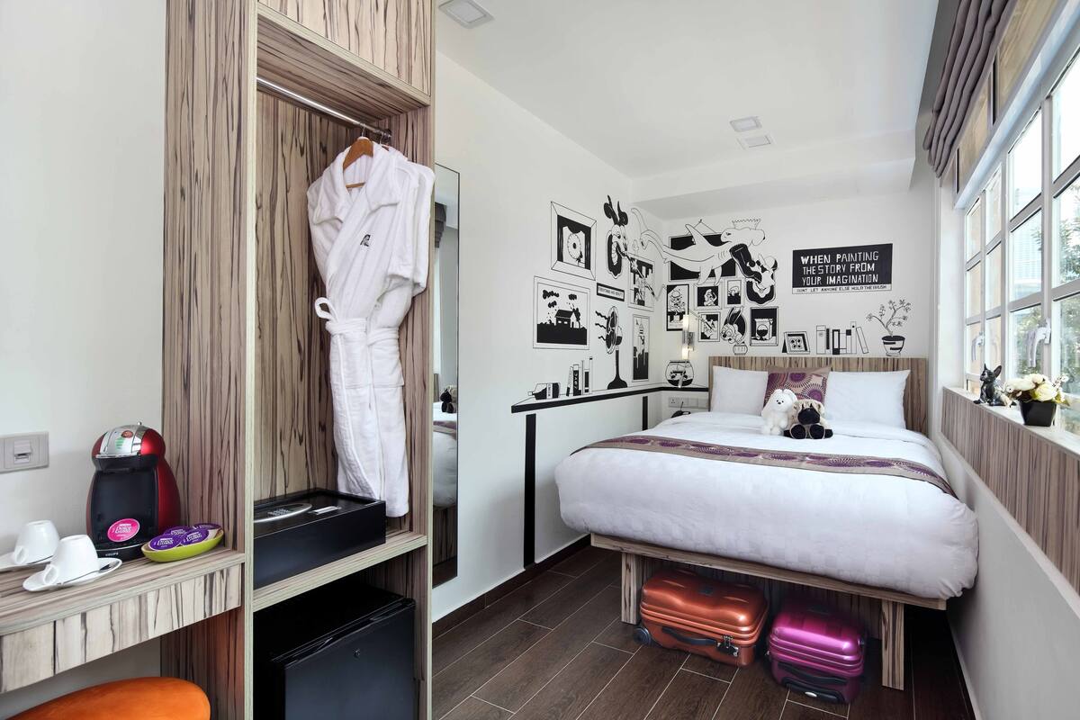 A hotel room with a bed, a wardrobe, a desk with a coffee maker, and a wall with black and white pictures.
