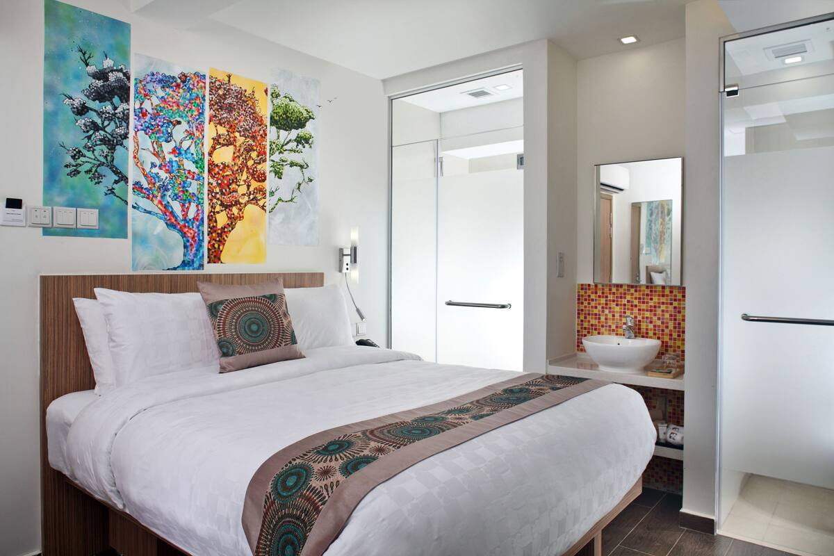 A modern hotel room with a large bed, a bathroom with a sink and mirror, and three colorful paintings on the wall.