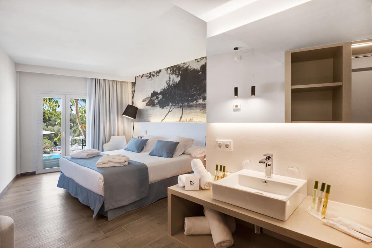 Une chambre d’hôtel moderne comprenant un lit, une salle de bain avec un lavabo et des articles de toilette, ainsi qu’une grande fenêtre donnant sur un paysage verdoyant.