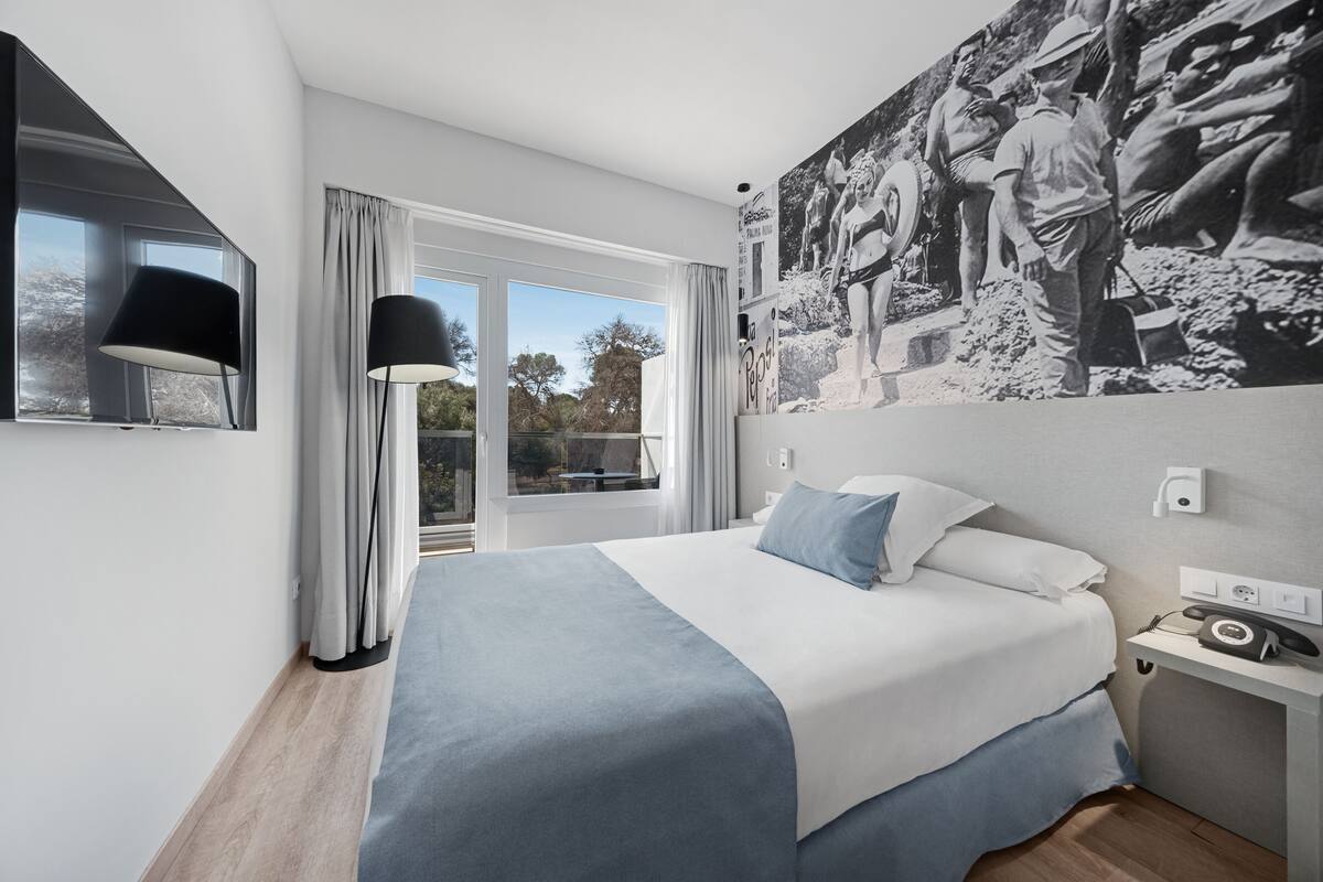 Une chambre d’hôtel moderne dotée d’un grand lit, d’un téléviseur à écran plat, d’une table de chevet avec un téléphone et d’une grande fenêtre donnant sur l’extérieur.