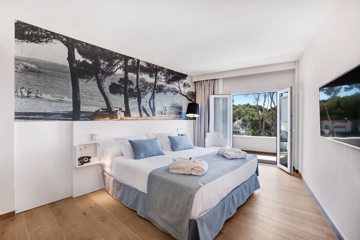 Une chambre d’hôtel moderne avec un grand lit, un tableau représentant une scène de plage, un balcon avec vue et une grande fenêtre.