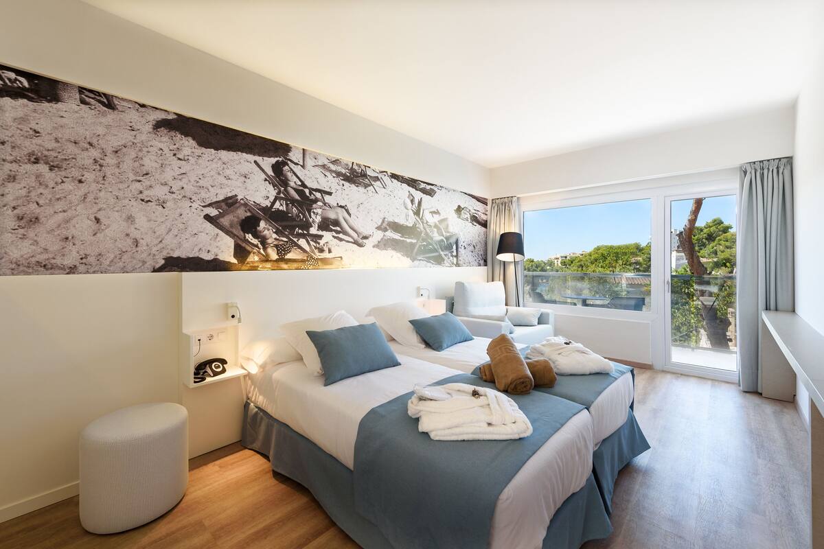Une chambre d’hôtel avec deux lits, une grande fenêtre donnant sur un beau paysage, un tableau au mur et une lampe.