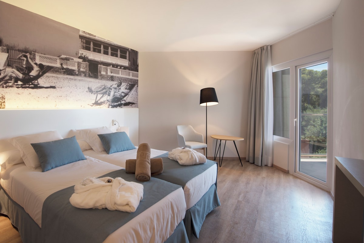 A modern hotel room with two beds, a large beach photo on the wall, a small table, and a view of the outdoors.