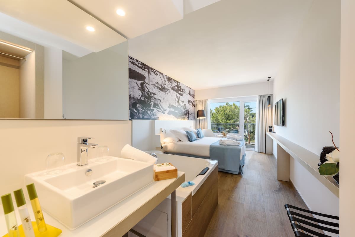 Une chambre d’hôtel moderne avec un grand lavabo blanc, un lit recouvert d’une couette bleue et blanche, un grand miroir et une vue sur l’extérieur.