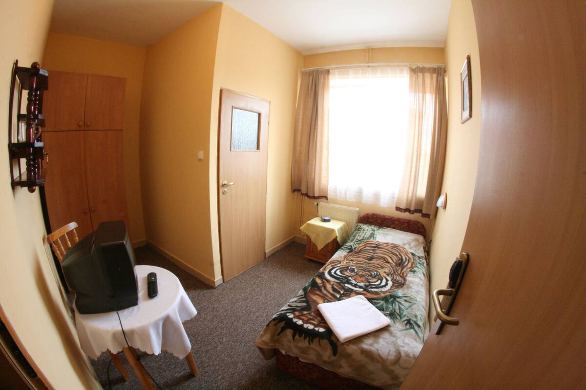 A small, compact room with a bed, a table with a chair, a wooden wardrobe, and a window with curtains.