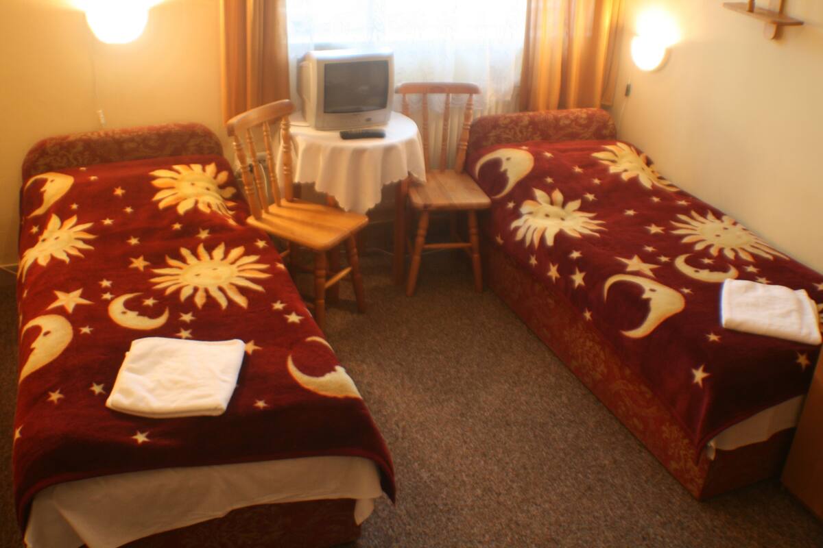 A room with two beds, each covered with a red blanket featuring moon and star patterns. There is a small table with a television and a chair in the center.