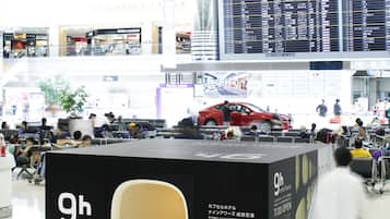 A modern airport terminal with a 9h kiosk, a digital flight information display, and a red car model.