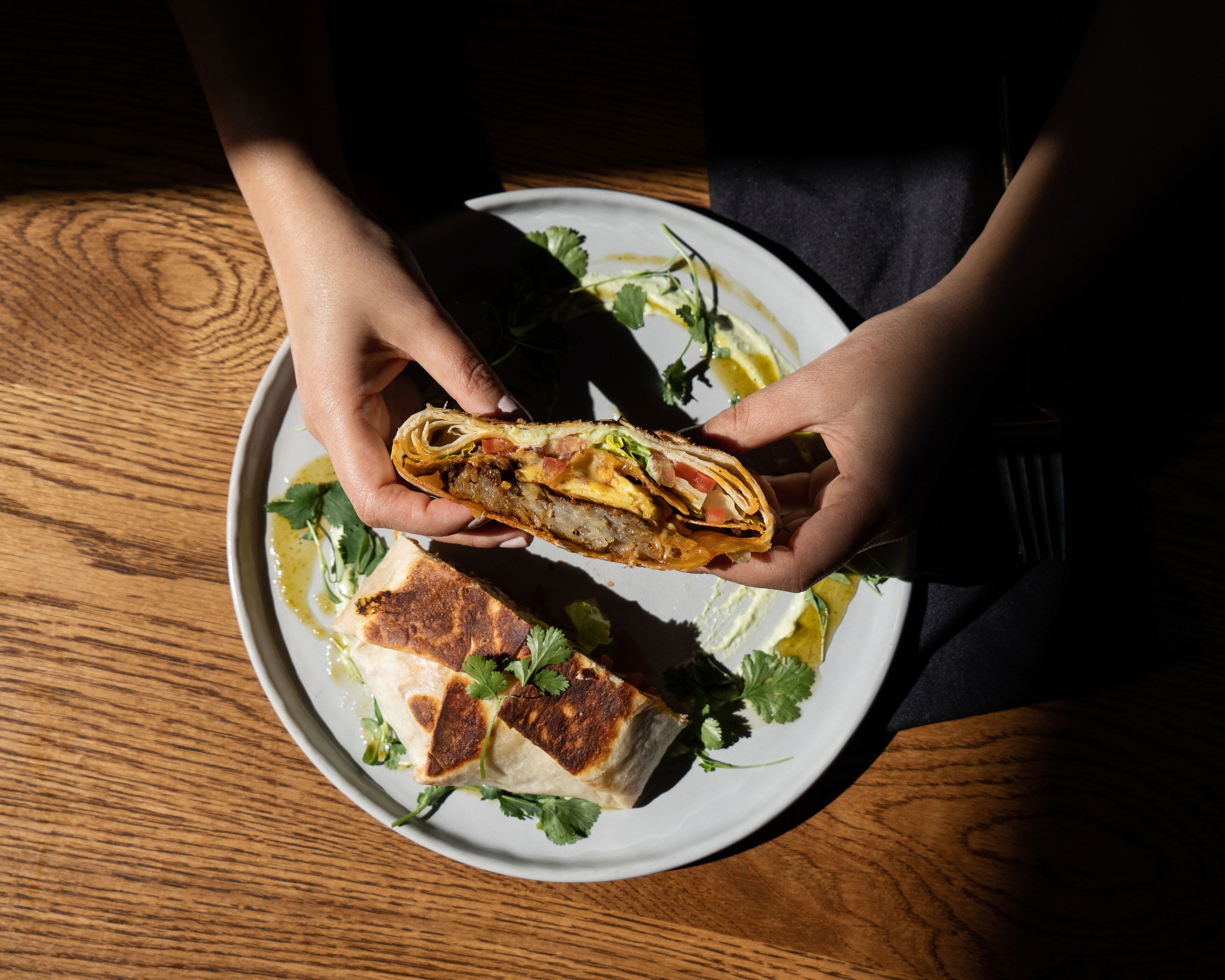 Una persona sosteniendo un burrito en un plato, con una mesa de madera al fondo.