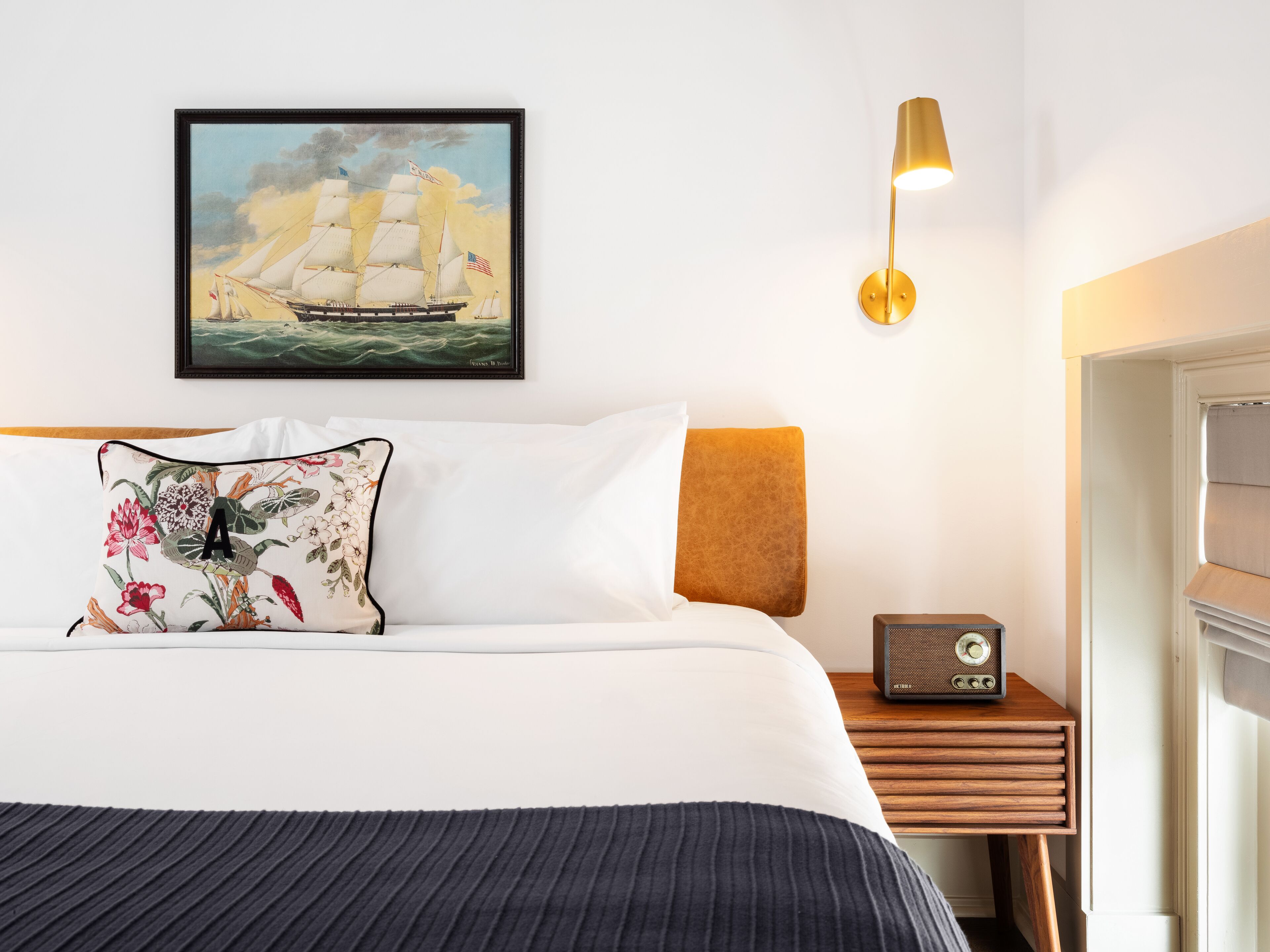 Ett välordnat hotellrum med en stor säng, en inramad tavla som föreställer ett segelfartyg, en sänglampa och en gammal radio placerad på ett träbord bredvid sängen.