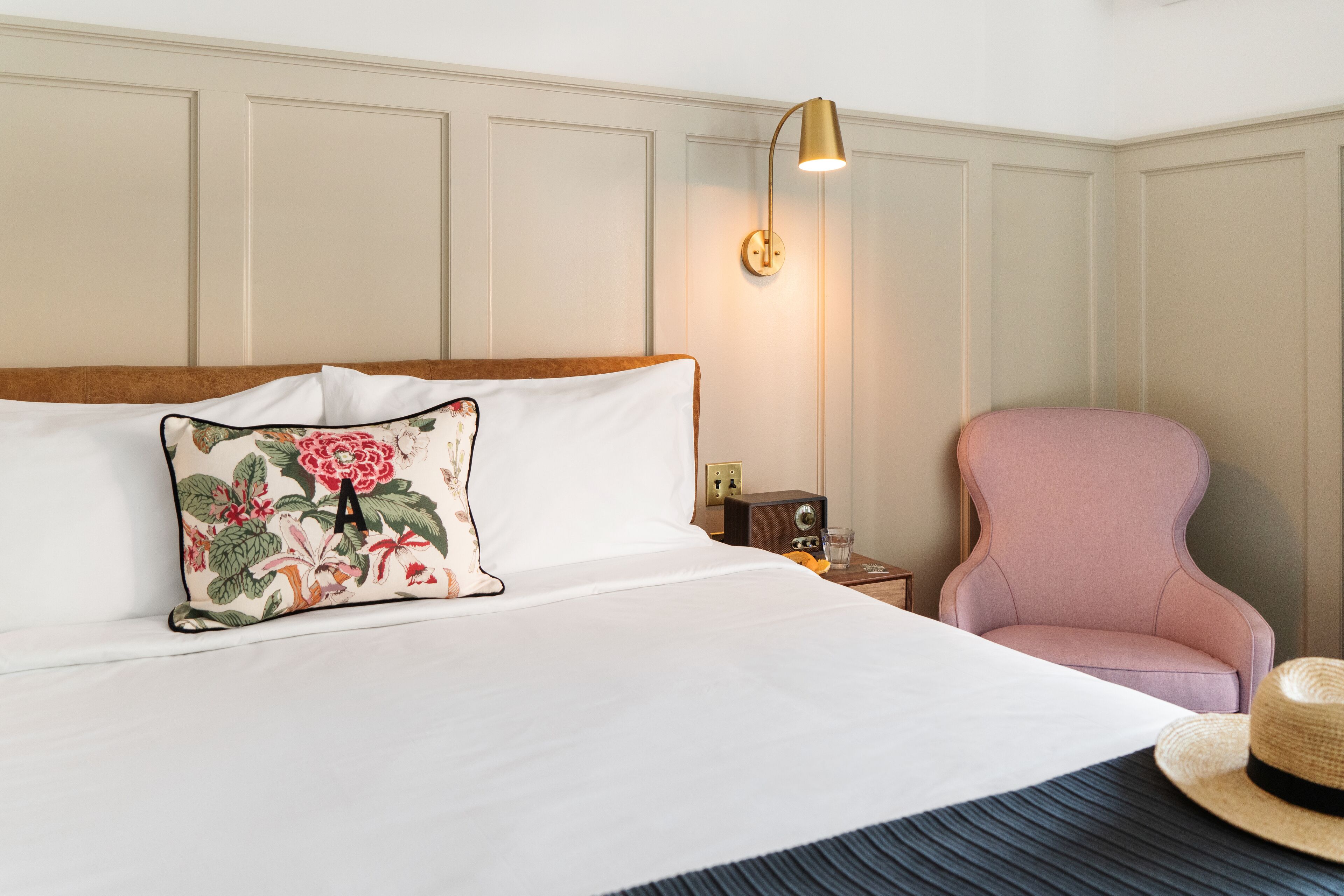 Una cama bien hecha con un cojín floral, una lámpara de noche, una silla rosa y un sombrero de paja en el suelo.