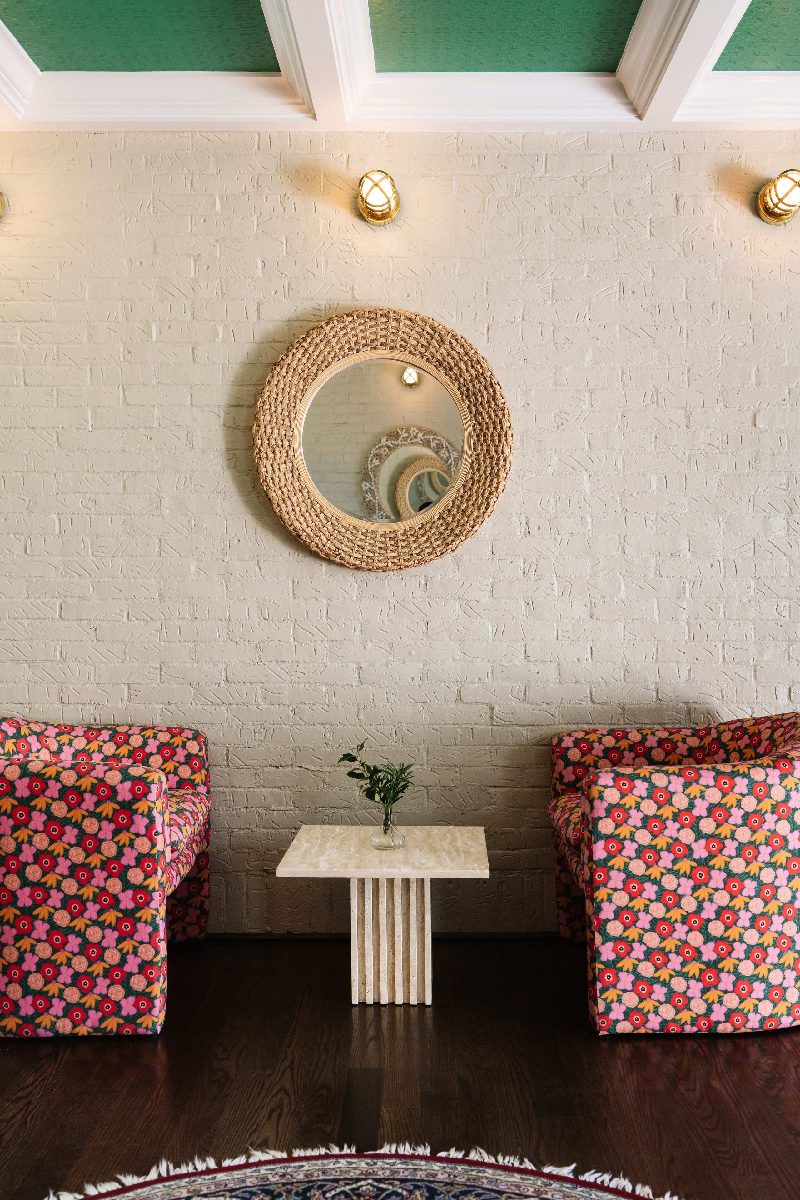 A room with a white brick wall, a round woven mirror, two floral-patterned sofas, a small white table with a plant, and a patterned rug.