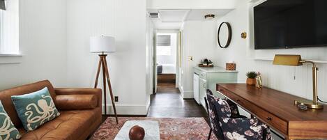 Een woonkamer met een bruine leren bank, een vloerlamp en een salontafel. Er is een keukenhoek met een houten bureau en een televisie.