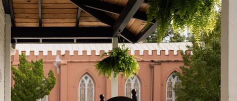 A covered porch with a wooden floor and a white railing. There are two rocking chairs and a hanging fern. In the background, there is a building with arched windows and a red facade.