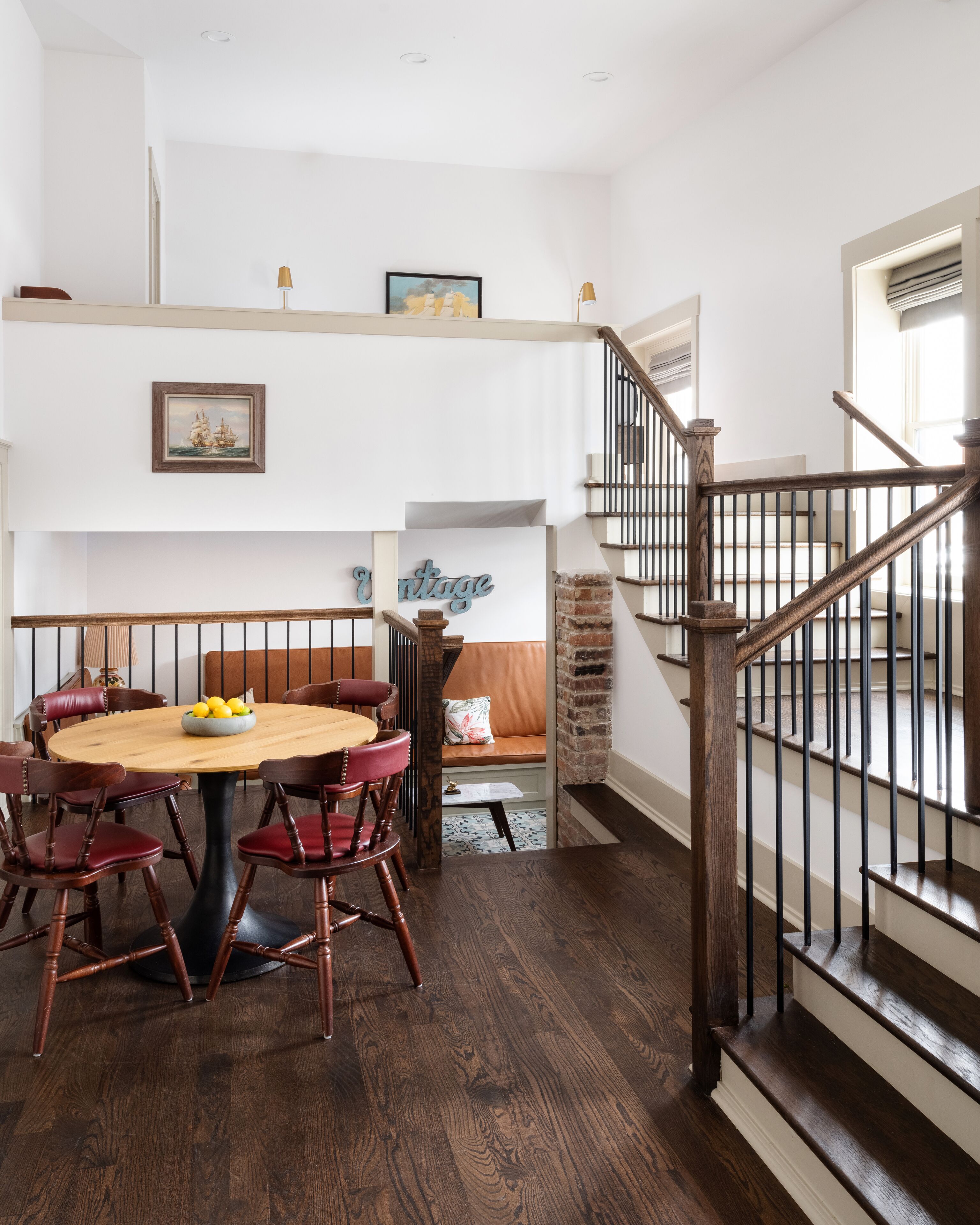 Un comedor con una mesa redonda de madera y sillas, una escalera con barandilla de madera, y un estante de pared con adornos.