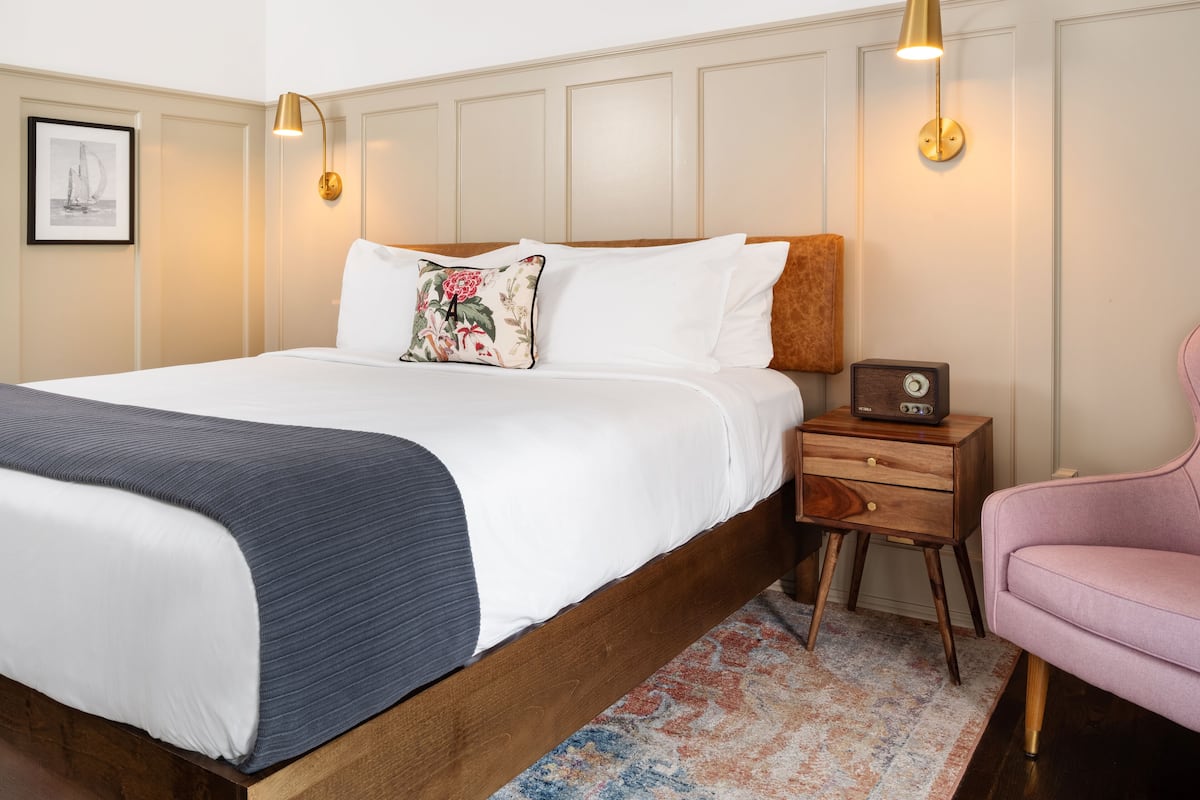 A neatly made bed with white linens, a floral pillow, and a dark throw blanket. There is a wooden nightstand with a radio and a pink chair to the right.