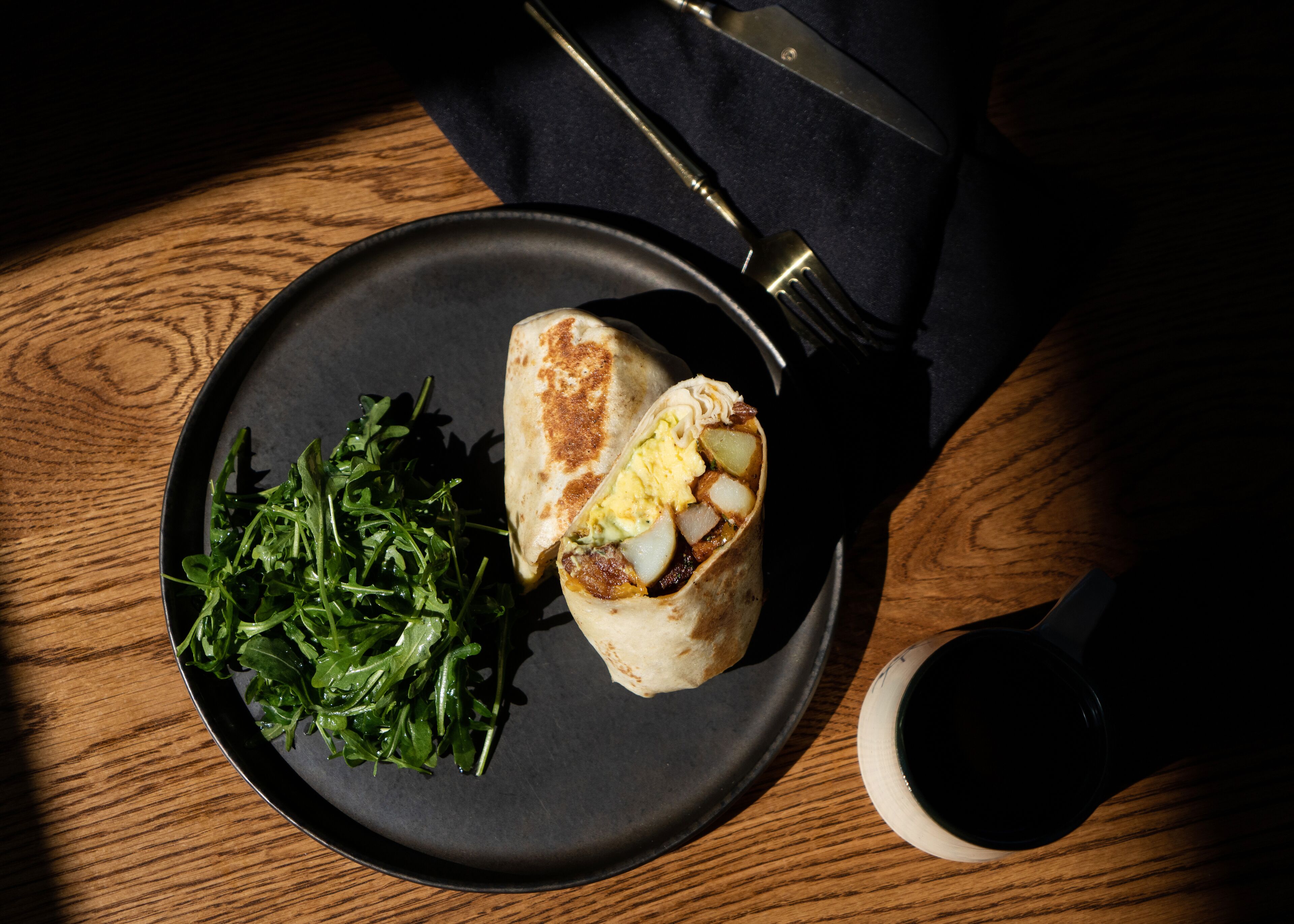 A plate with a wrap, greens, and a cup on a wooden table.