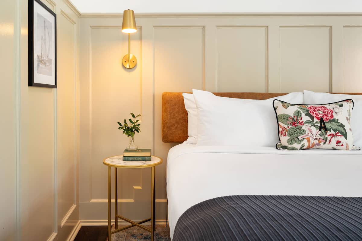 A neatly arranged hotel room with a bed, a bedside table with a lamp, a framed picture on the wall, and a small plant.