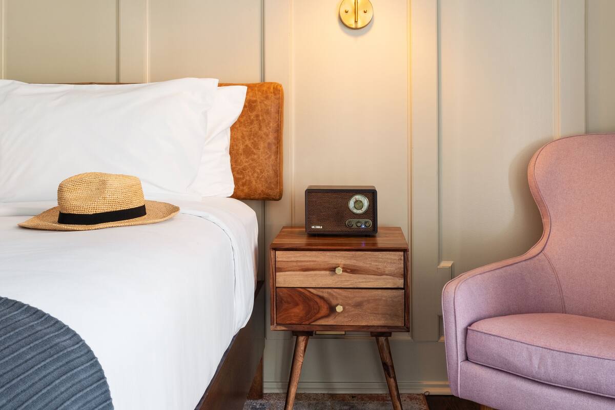 A neatly arranged hotel room with a bed, a bedside table with a radio, a pink armchair, and a patterned rug.