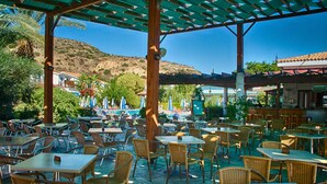 An outdoor café with tables and chairs, a pool, and a mountainous backdrop. - Hylatio Tourist Village (Pissouri)