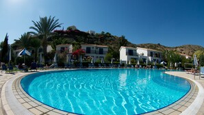 A large, clear swimming pool with lounge chairs and umbrellas around it, set against a backdrop of buildings and hills. - Hylatio Tourist Village (Pissouri)