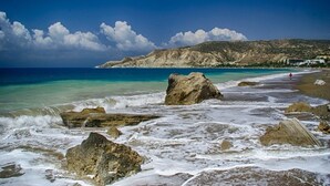 A beach with large rocks and waves, a clear sky, and a distant hill with buildings. - Hylatio Tourist Village (Pissouri)