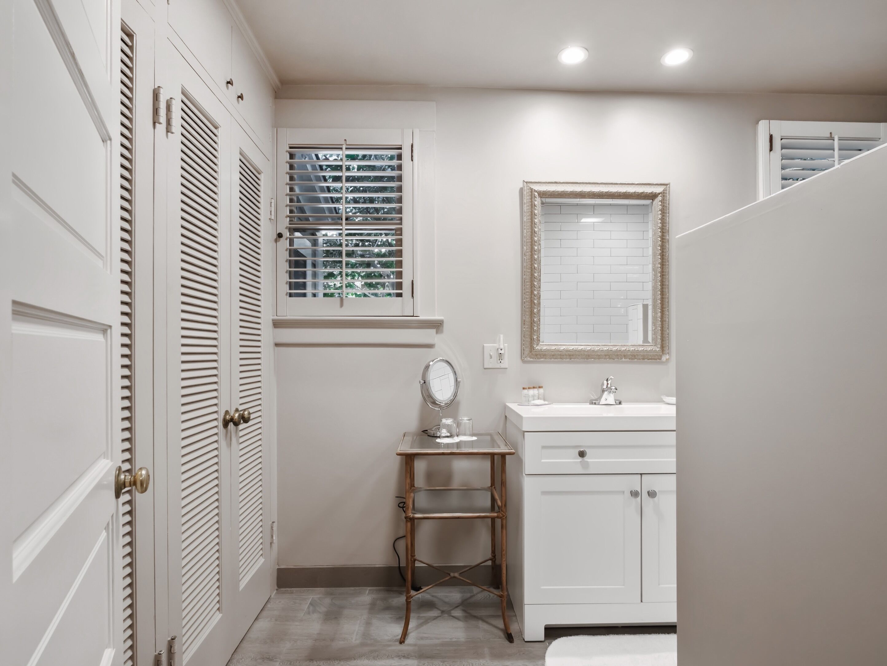 A bathroom with a vanity, a mirror, a window with blinds, and a door with louvered panels.