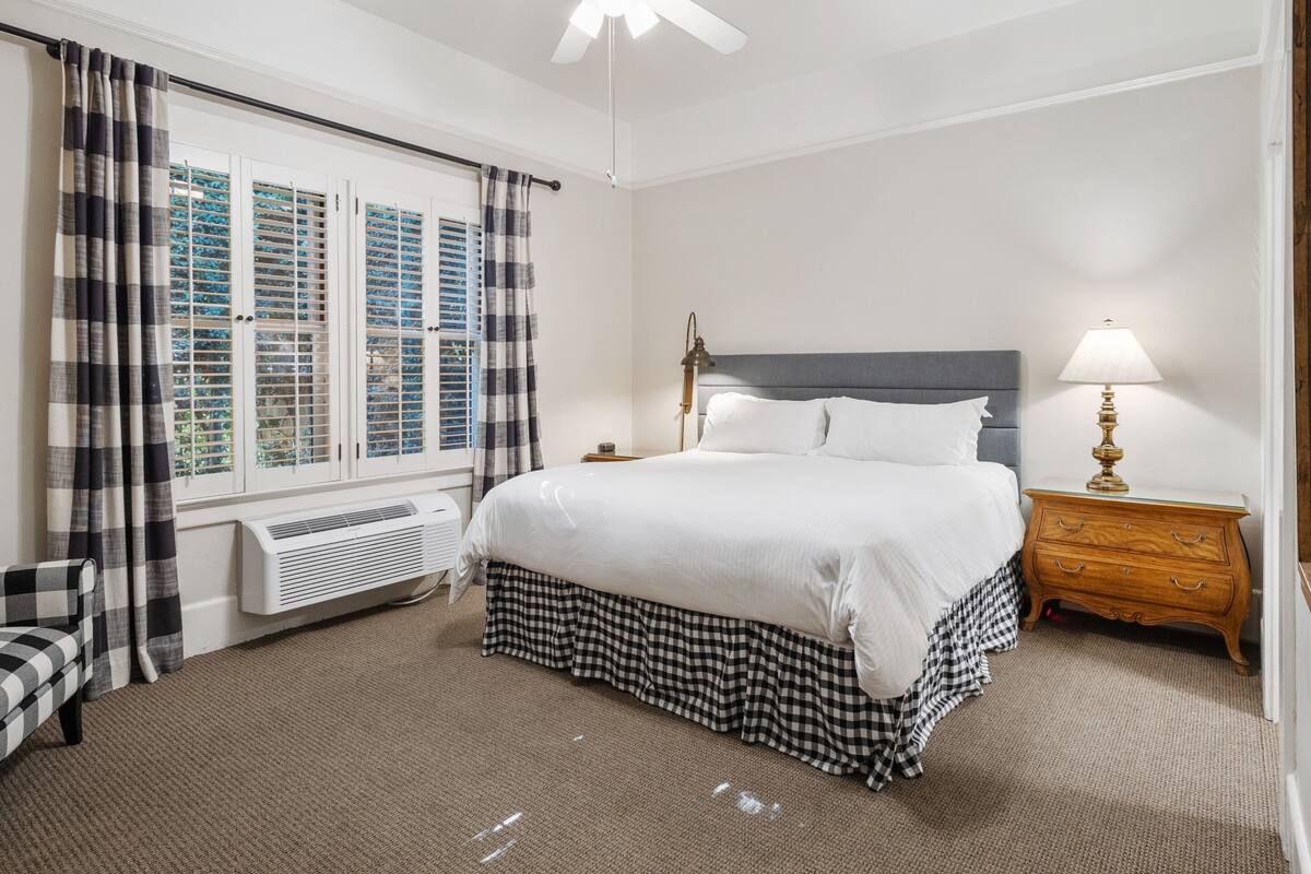 Une chambre d’hôtel comprenant un lit, une table de chevet, une lampe, une fenêtre avec des rideaux, un climatiseur et un ventilateur de plafond.
