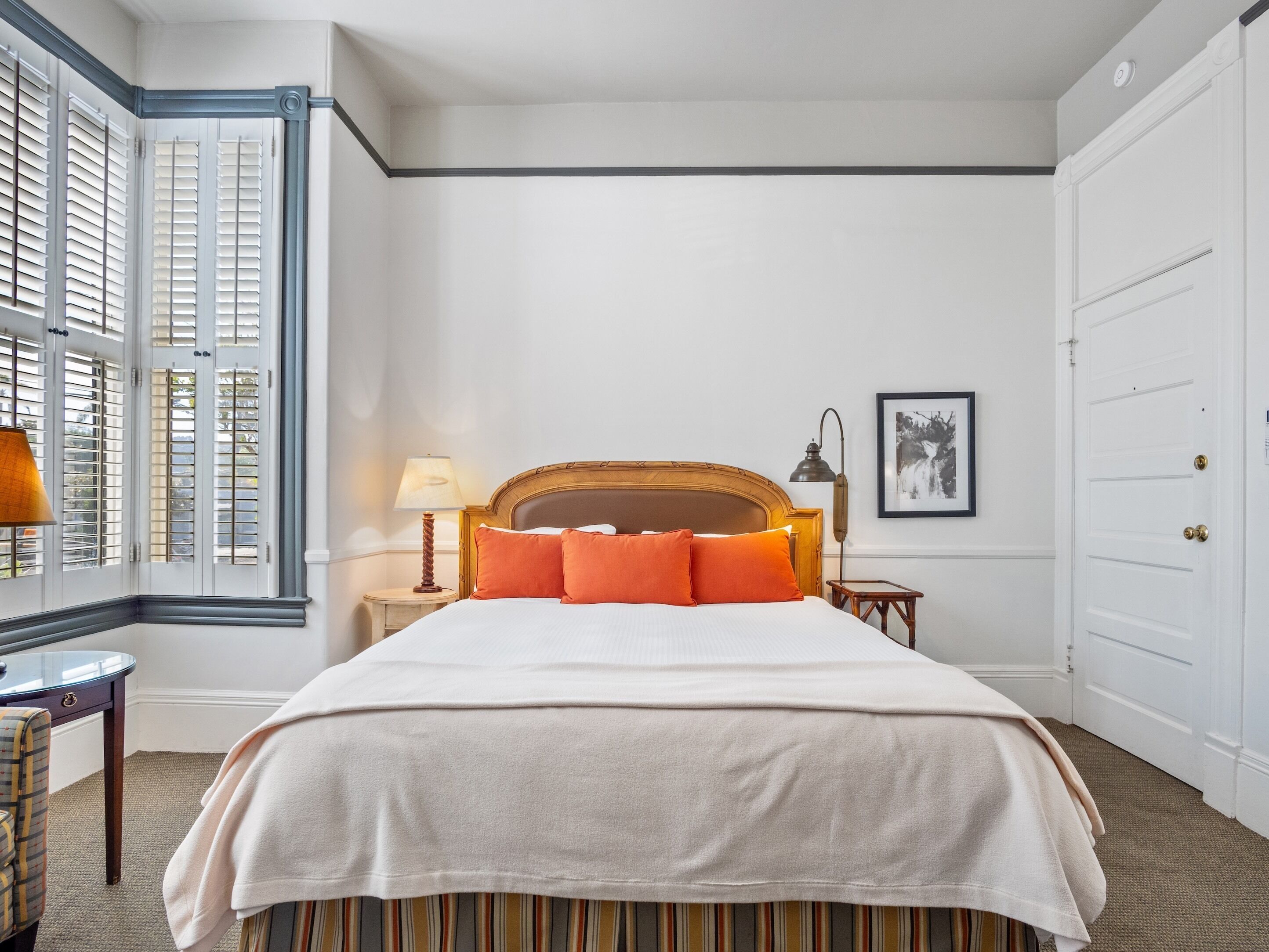 Een netjes ingerichte slaapkamer met een groot bed, een nachtkastje met een lamp, een lijst met een afbeelding aan de muur en een raam met luiken.