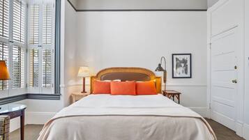 Een netjes ingerichte slaapkamer met een groot bed, een nachtkastje met een lamp, een lijst met een afbeelding aan de muur en een raam met luiken.