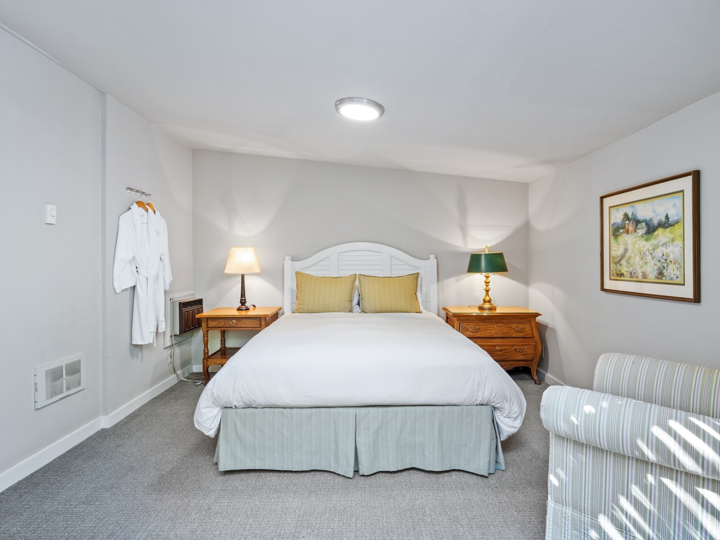 A neatly arranged bedroom with a large bed, two bedside tables with lamps, a chair, and a painting on the wall.