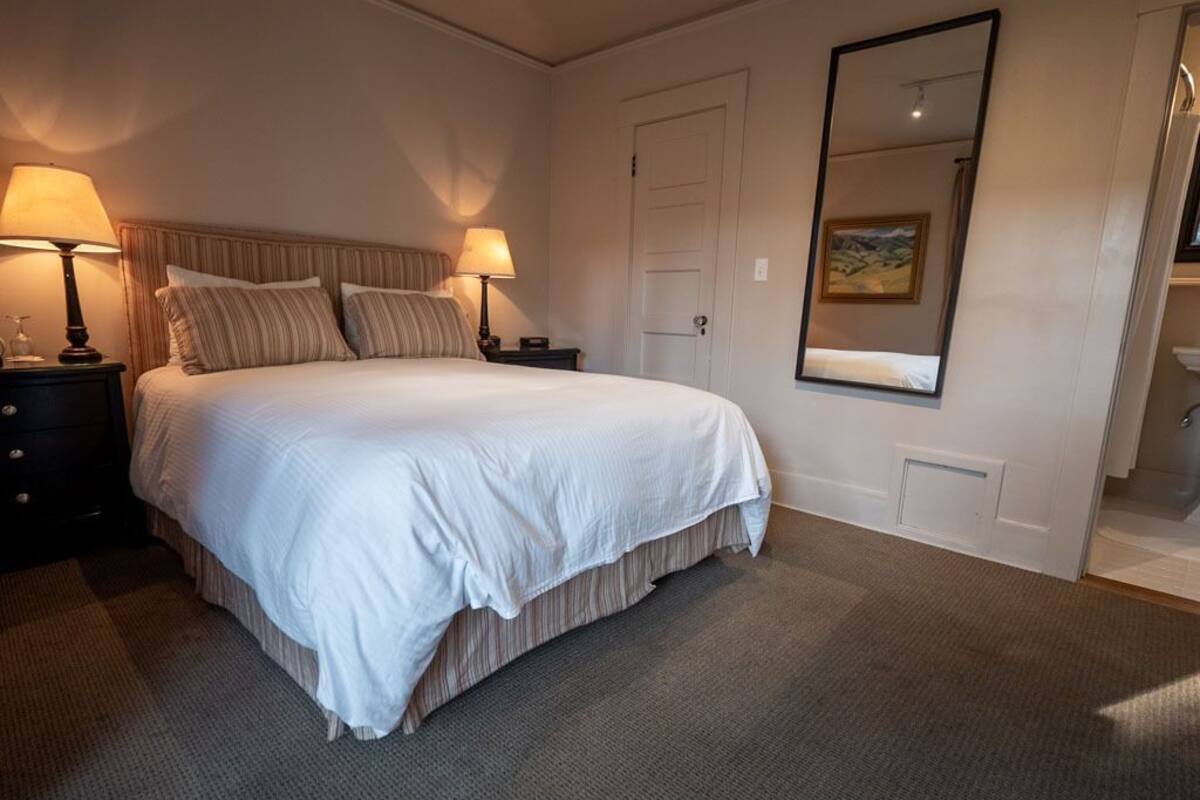 Un lit bien fait, avec une couette blanche et des oreillers à rayures, deux lampes de chevet, une porte et un grand miroir au mur.