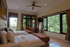 Habitación de hotel con dos camas, ventilador de techo, televisión y ventanas amplias que dan a zonas verdes.