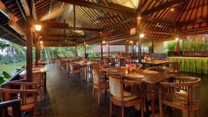 An indoor dining area with wooden tables and chairs, a high ceiling with wooden beams, and a view of greenery outside. - Cempaka Belimbing Villas (Pupuan)