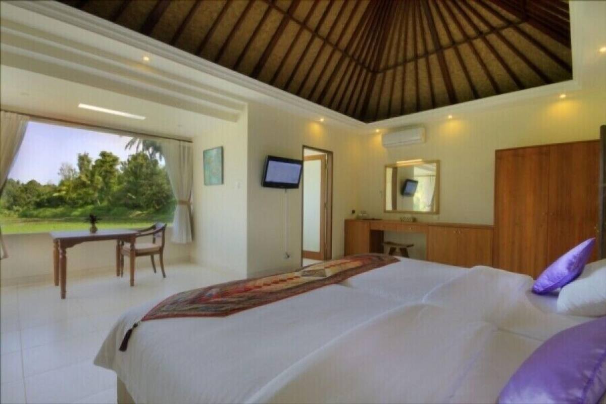 Een slaapkamer met een groot bed, een bureau en een stoel. Er is een televisie aan de muur en een raam met uitzicht op groen.
