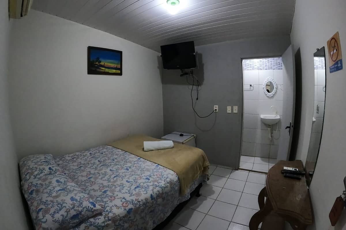 A small, simple room with a bed, a TV mounted on the wall, a bathroom with a sink and mirror, and a door with a 'No Smoking' sign.