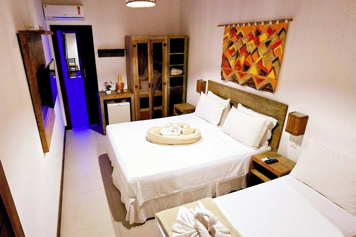 A hotel room with a bed, a bedside table, a nightstand, a wardrobe, and a bathroom visible through an open door.