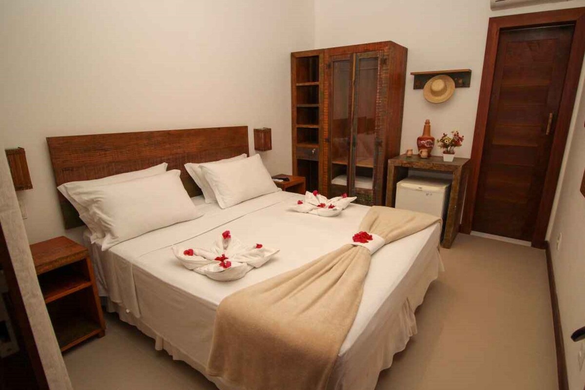 Una cama bien hecha con sábanas blancas y un plaid color beige, un armario de madera y una mesita de noche con una lámpara.